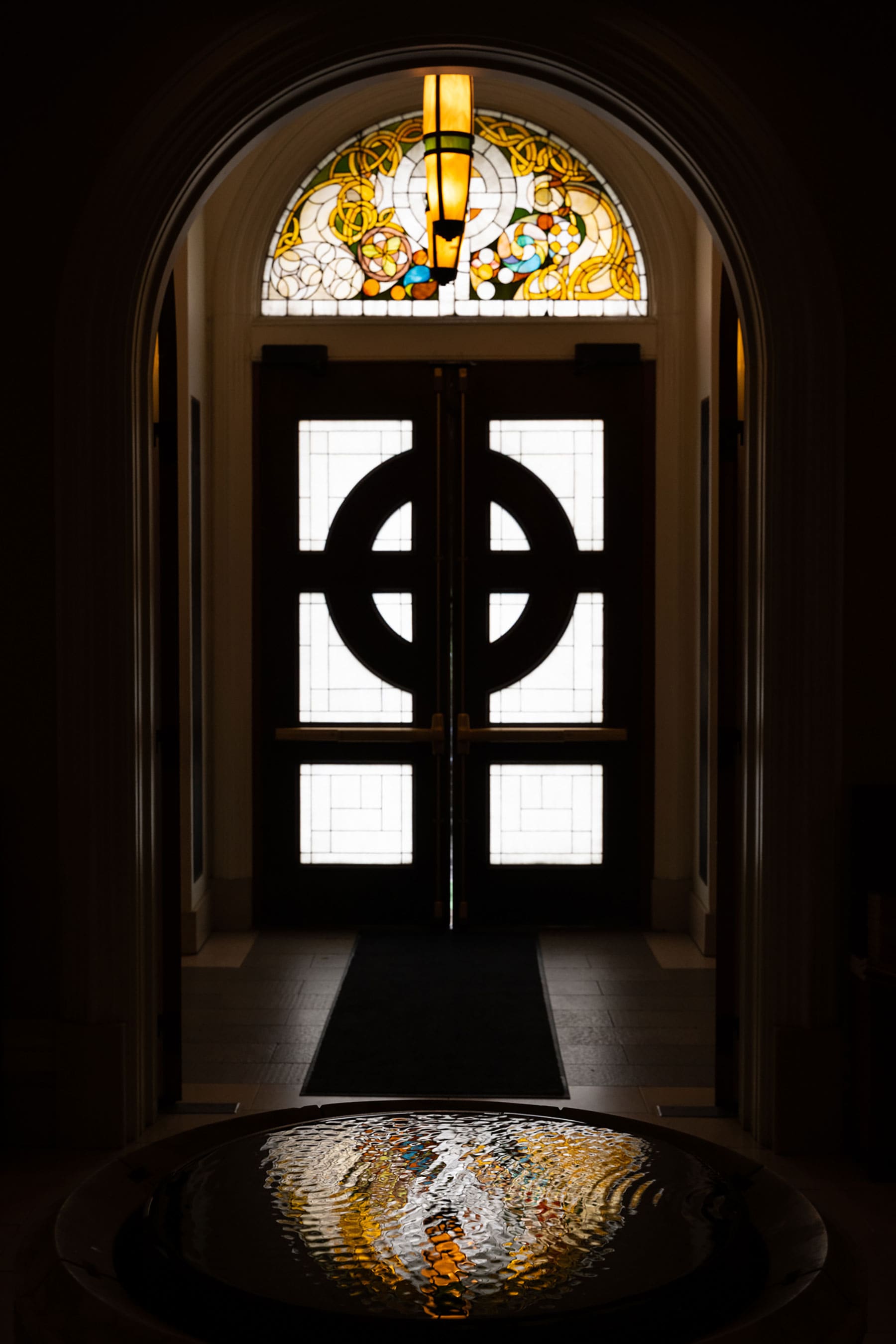 The image shows an arched entryway with dark double doors featuring geometric glass cutouts, a colorful semicircular stained-glass transom window above, and a glowing pendant light fixture