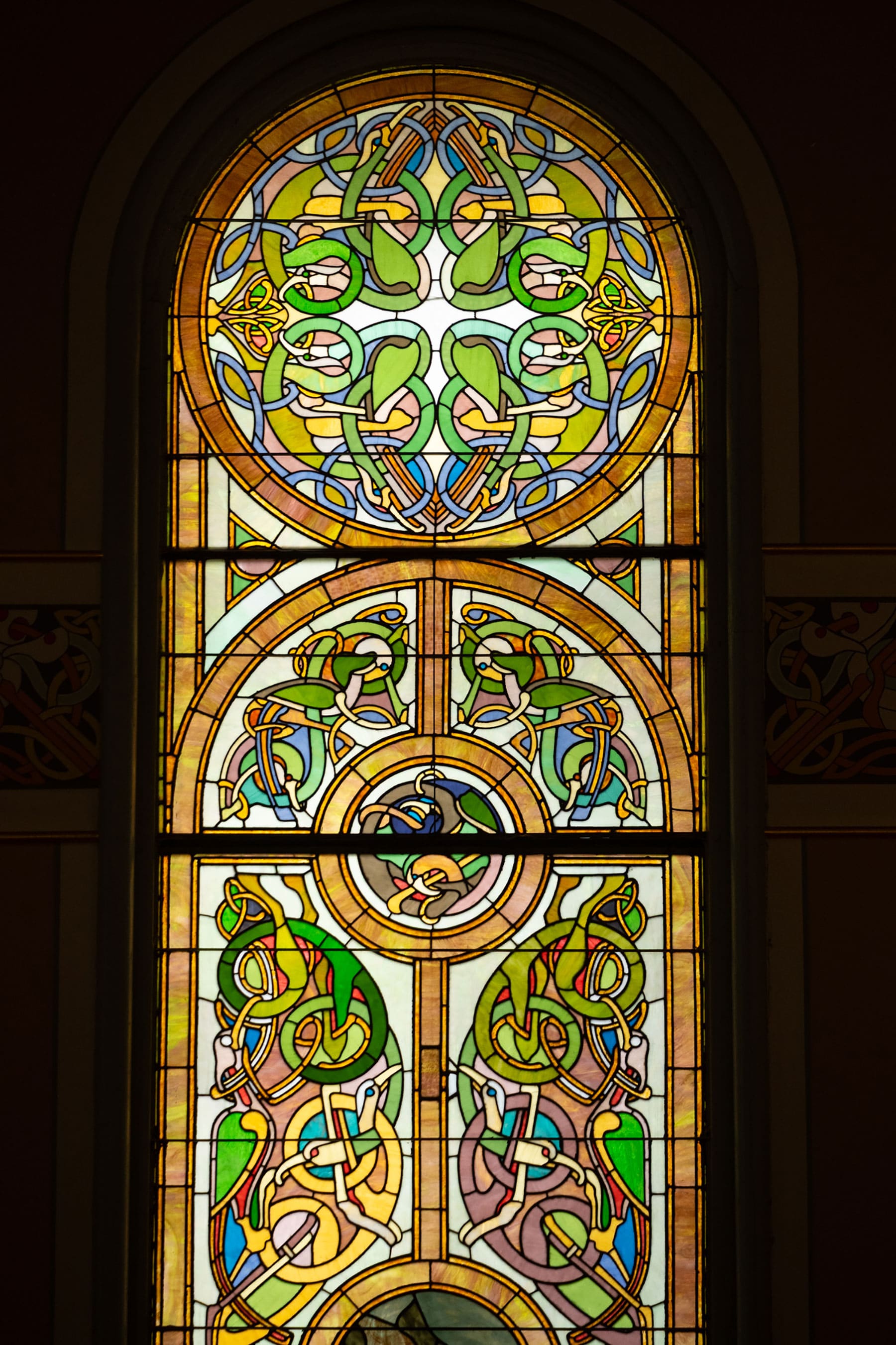 An arched, vertical stained glass window featuring intricate Celtic knotwork and zoomorphic designs in shades of green, gold, blue, and cream.