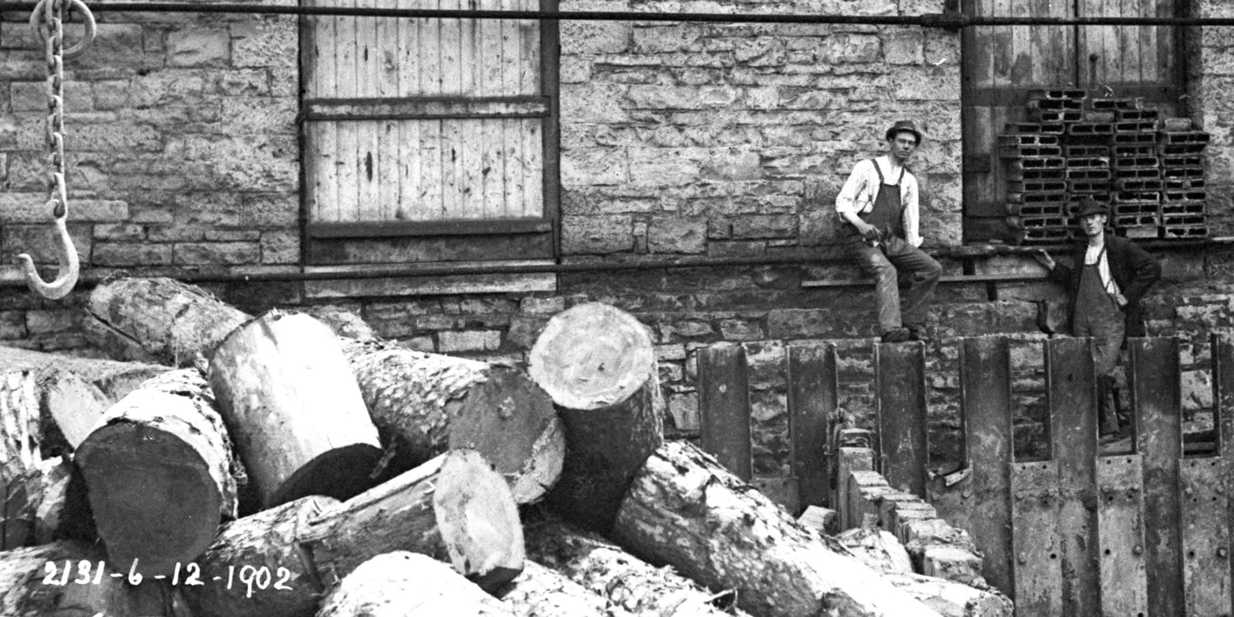 Industrial workers posing next to stacked timber logs and metal sheeting near a stone building from 1912