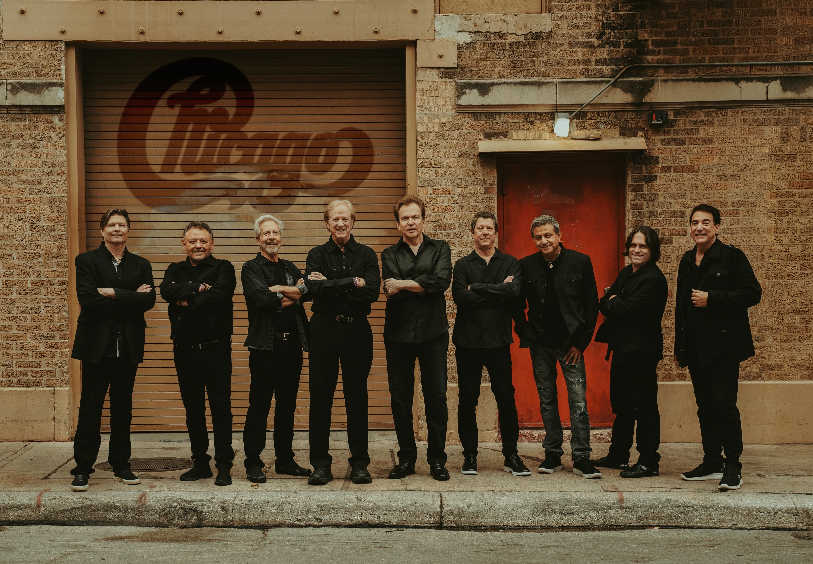 The members of the band Chicago pose in front of a brick building with the Chicago logo on it