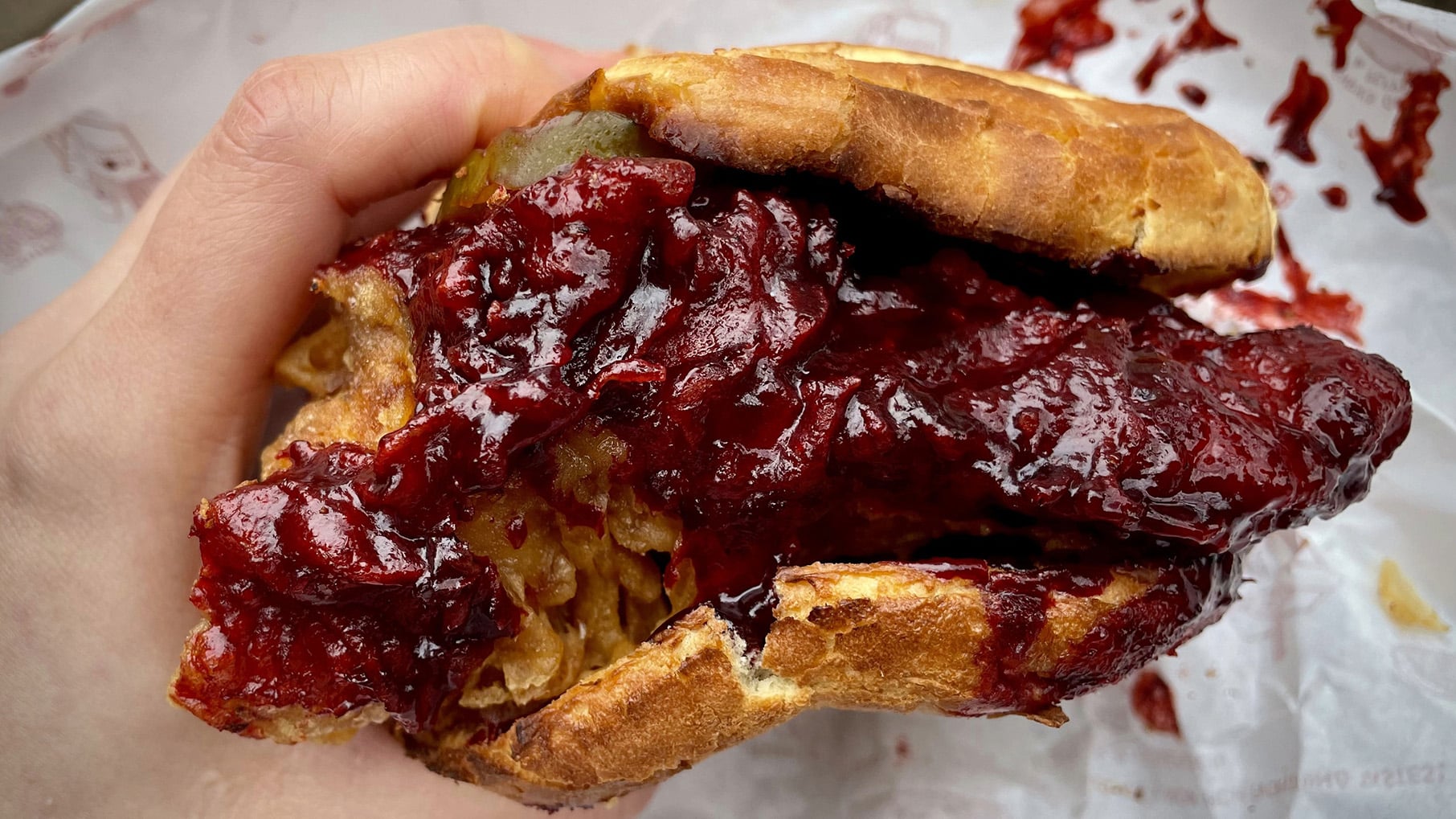 A hand holds a fried chicken sandwich soaked in red sauce