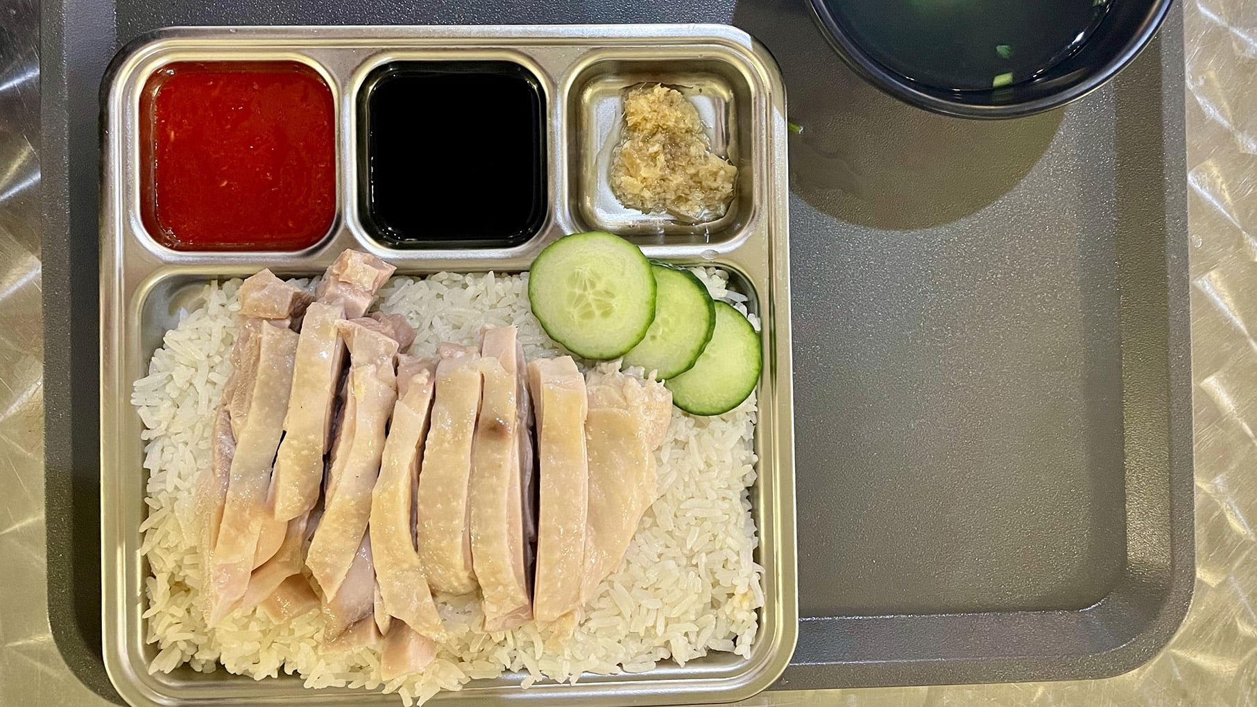 A tray with chicken on rice along with some sauces and pickles