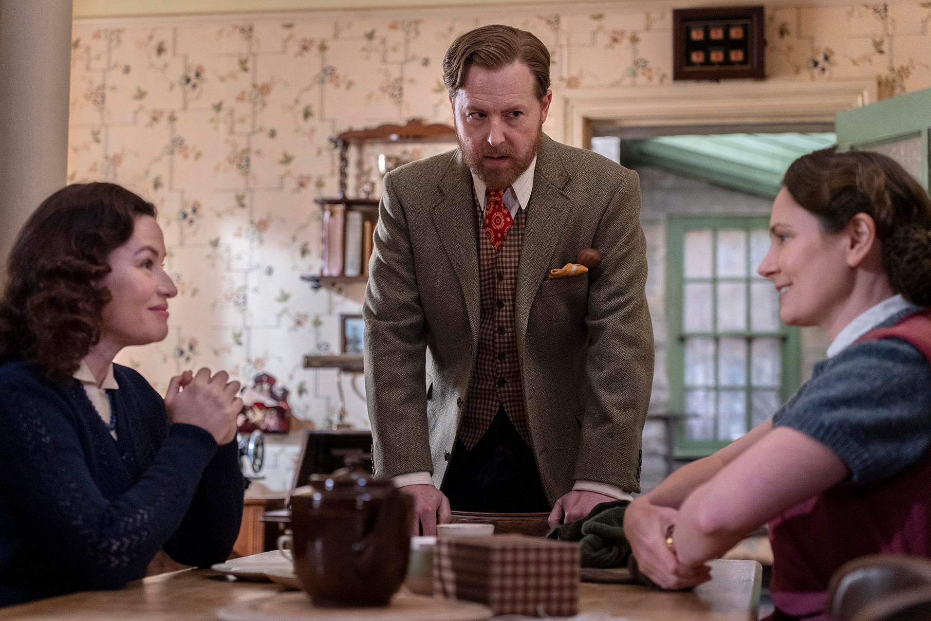 Dorothy and Mrs. Hall face each other at the kitchen table while Siegfried stands between them