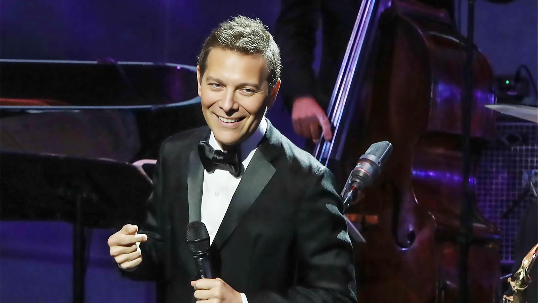 Michael Feinstein holds a microphone onstage in a tuxedo
