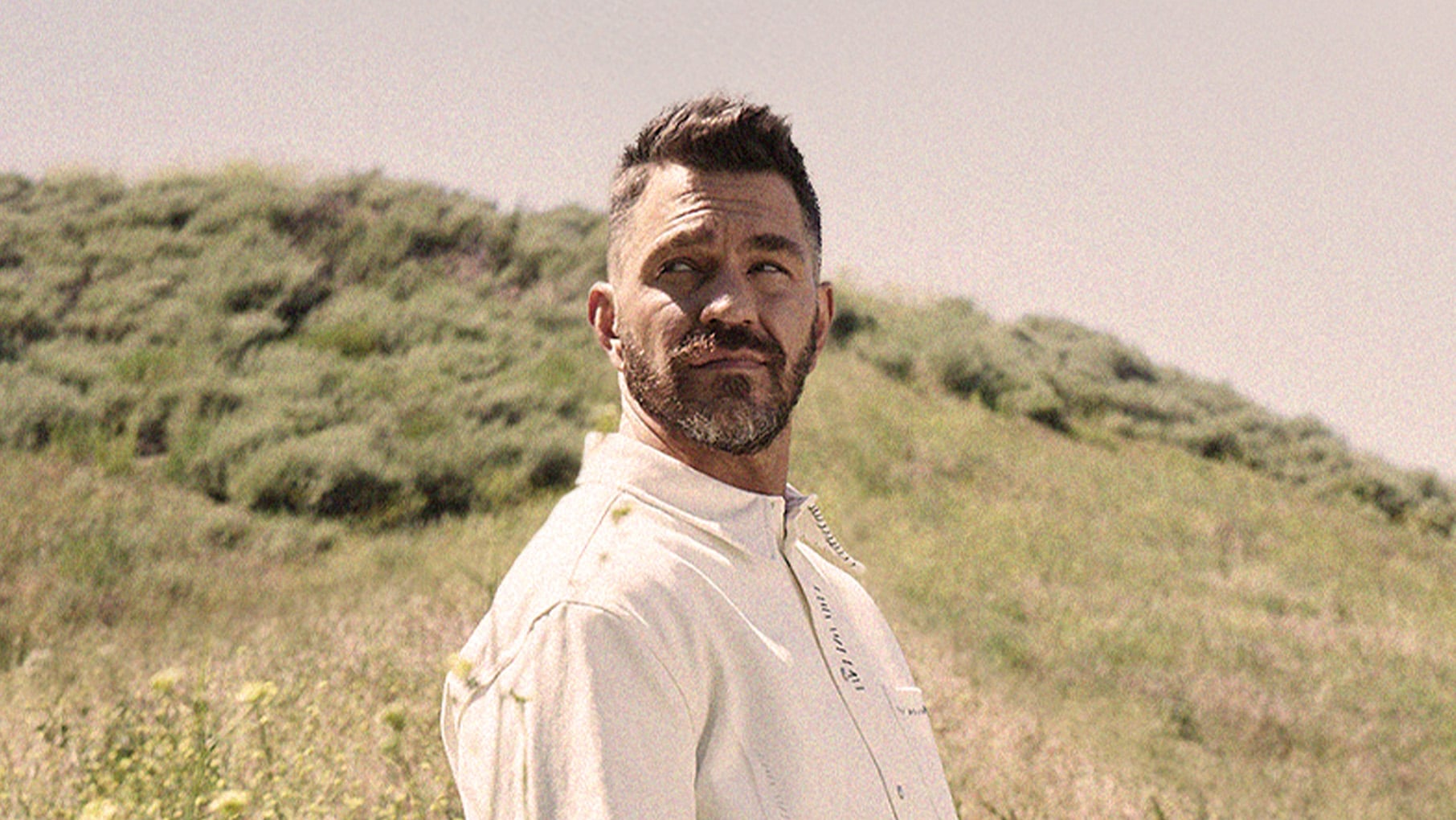 Andy Grammer stands outside in front of a hill in white