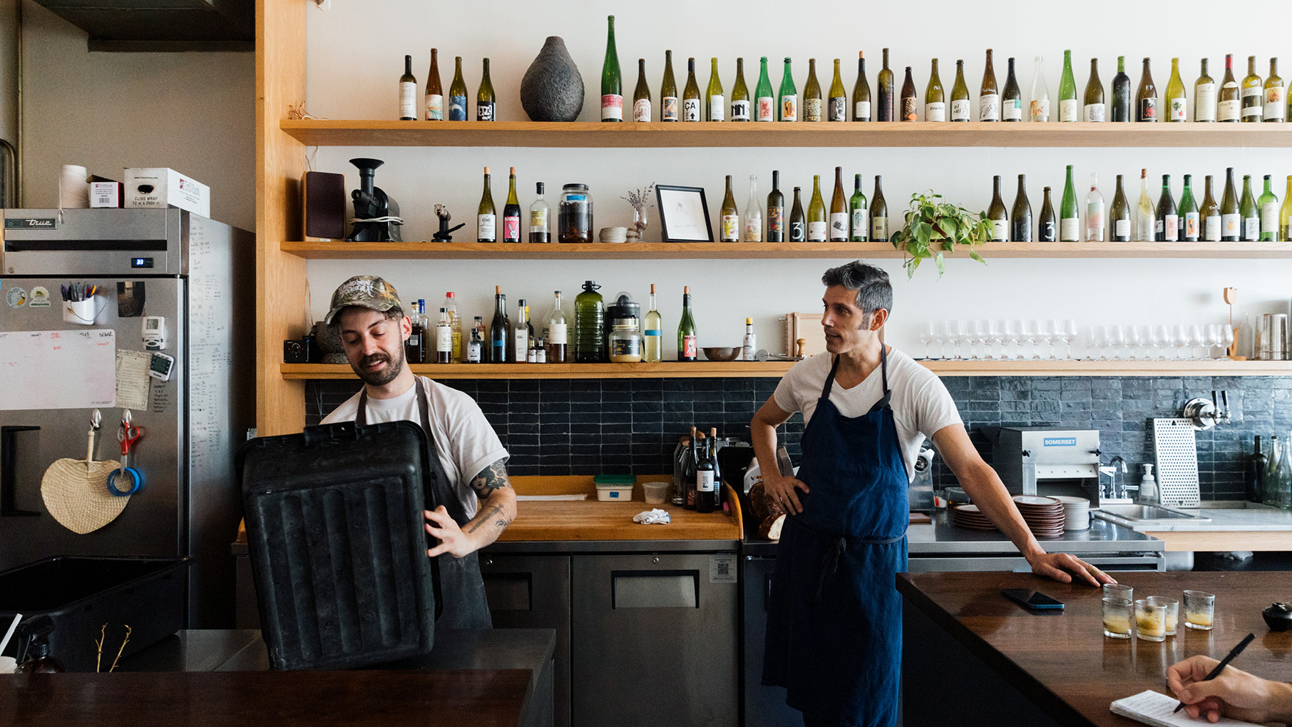 A man in a baseball cap empties a bin while another man in an apron watches, in front of a wall of bottles