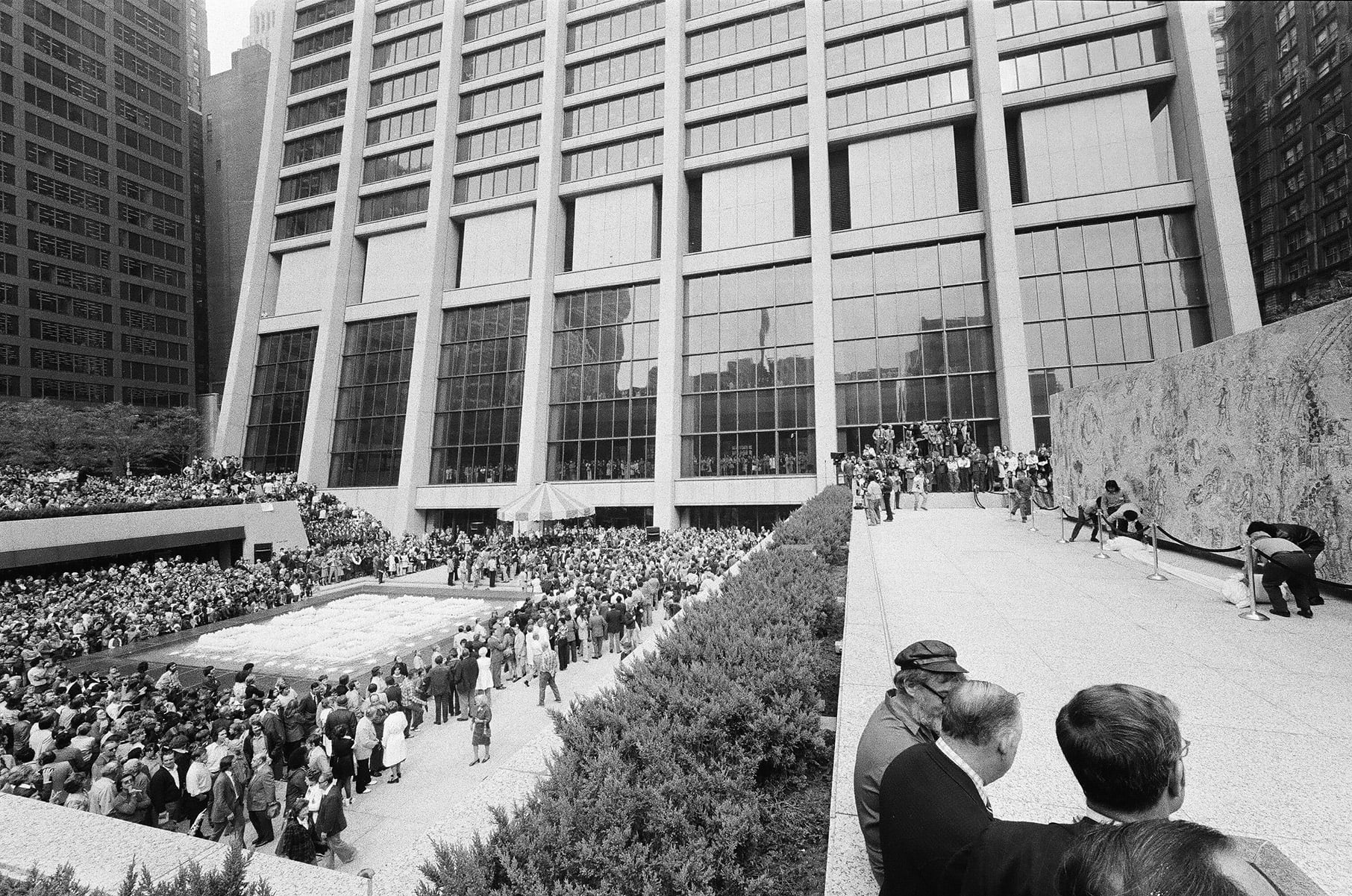 A black and white photo of a crowd of people in a plaza under a curving skyscraper, with Marc Chagall's The Four Seasons mosaic on the right