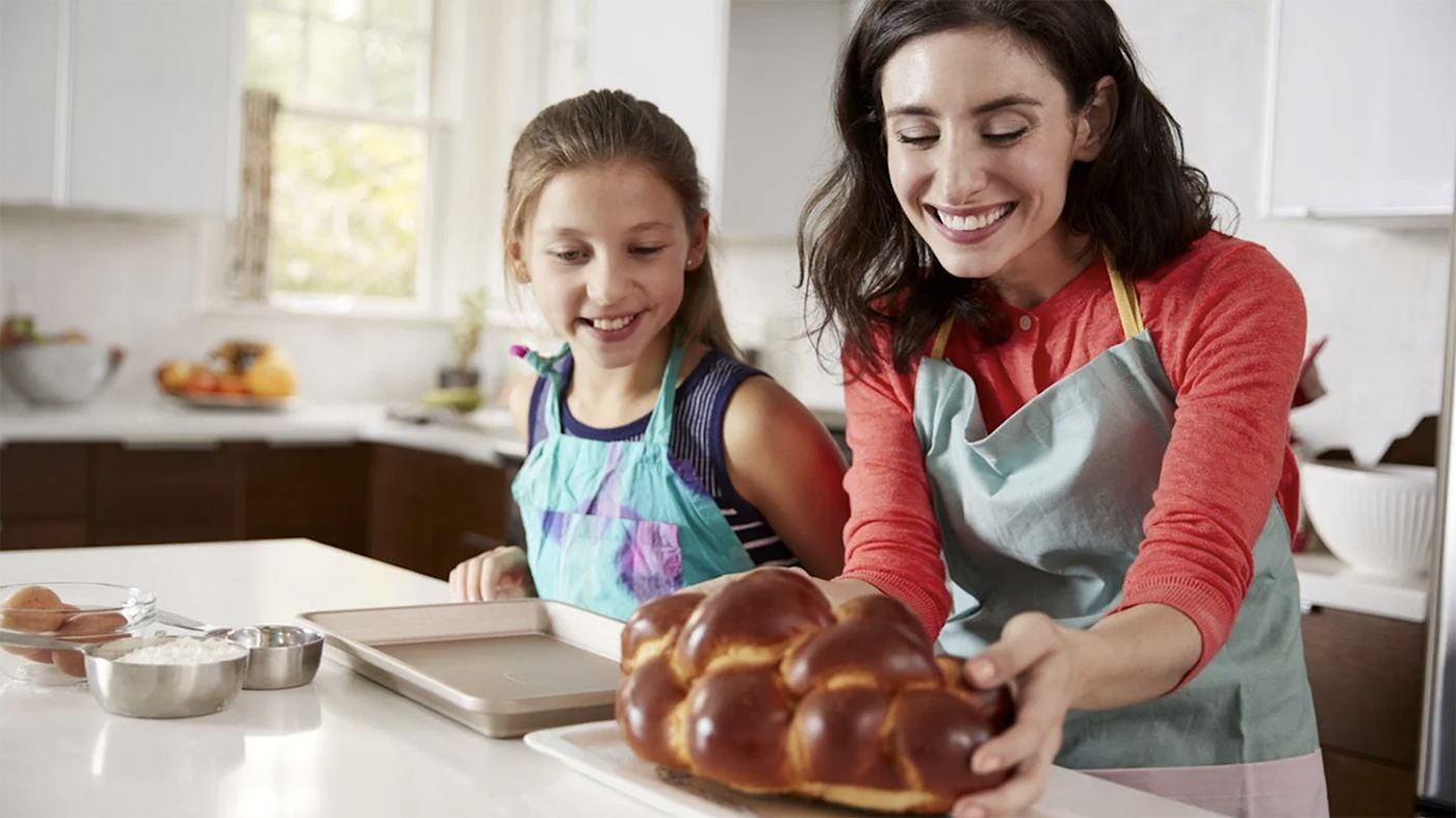 A woman and girl smiling while lifting challah off a baking tray