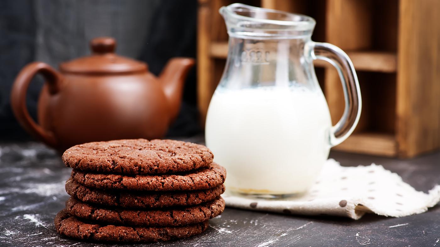 Five chocolate ginger molasses cookies stacked on top one another, with a pitcher of milk and a brown teapot in the backdrop