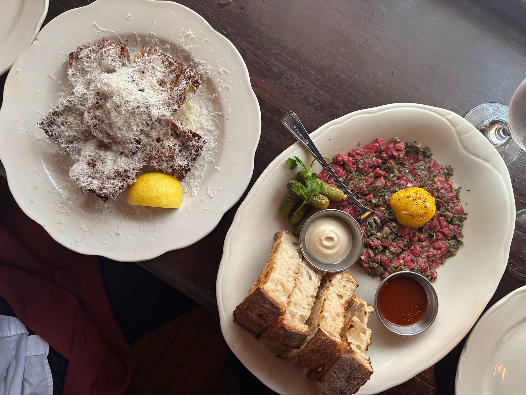 Artichoke showered in cheese on a plate next to a plate of tartare with an egg yolk, bread, a tin of sauce, and gherkins