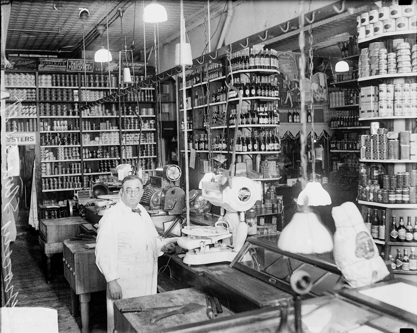 A black and white photo of a clerk behind a counter in a grocery store stocked with bottles and cans on shelves