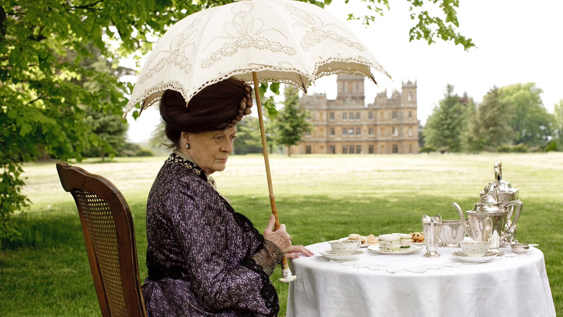 Violet sits under a parasol at a table set up outside Downton Abbey