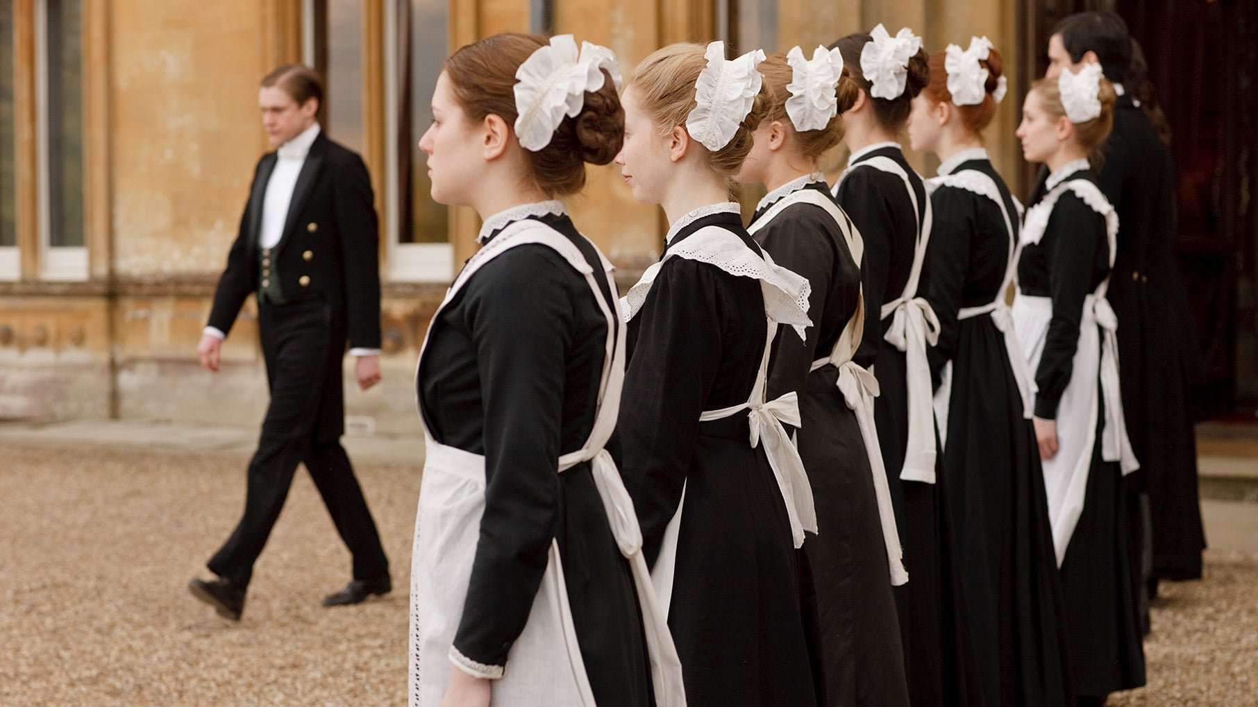 A man in a tuxedo walks down a line of maids outside