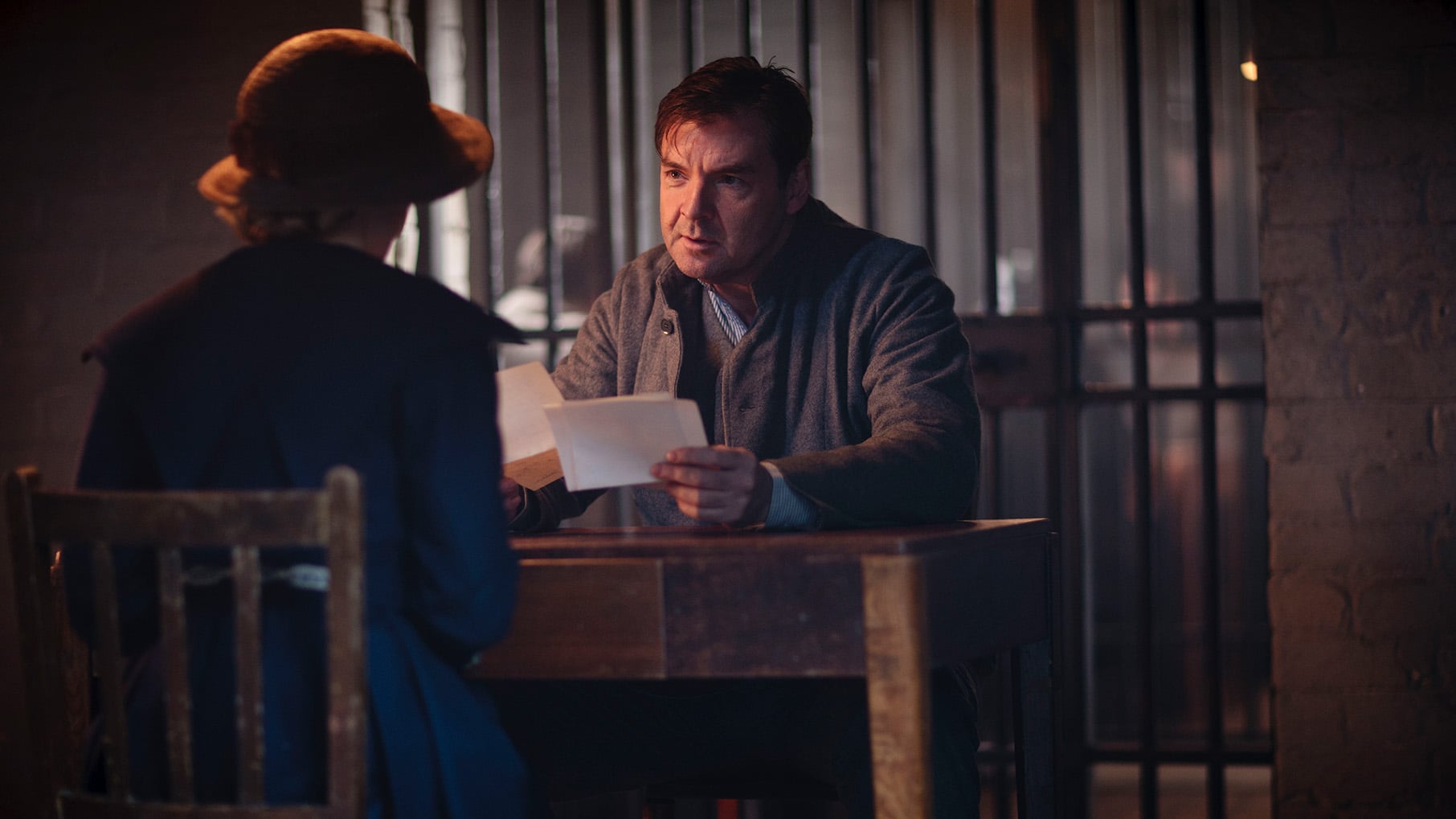 Bates holds a paper and sits across a desk from Anna in a prison cell