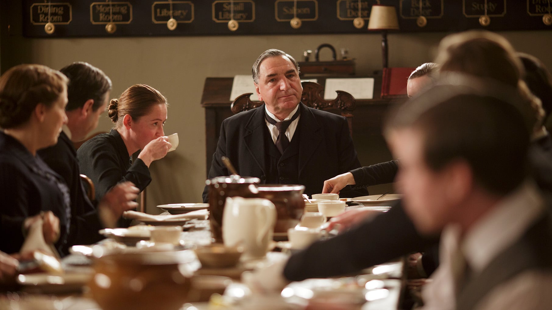 Carson sits at the head of a table full of servants eating, with coffee cups set out