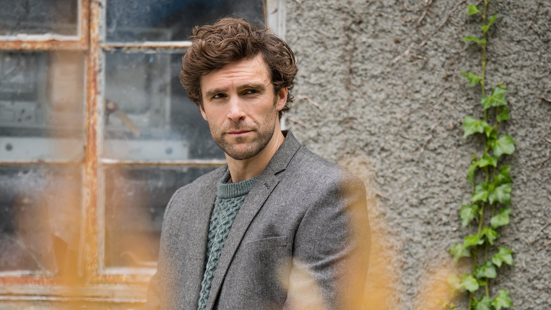 Ewan stands outside a window in a sweater, jacket, and kilt
