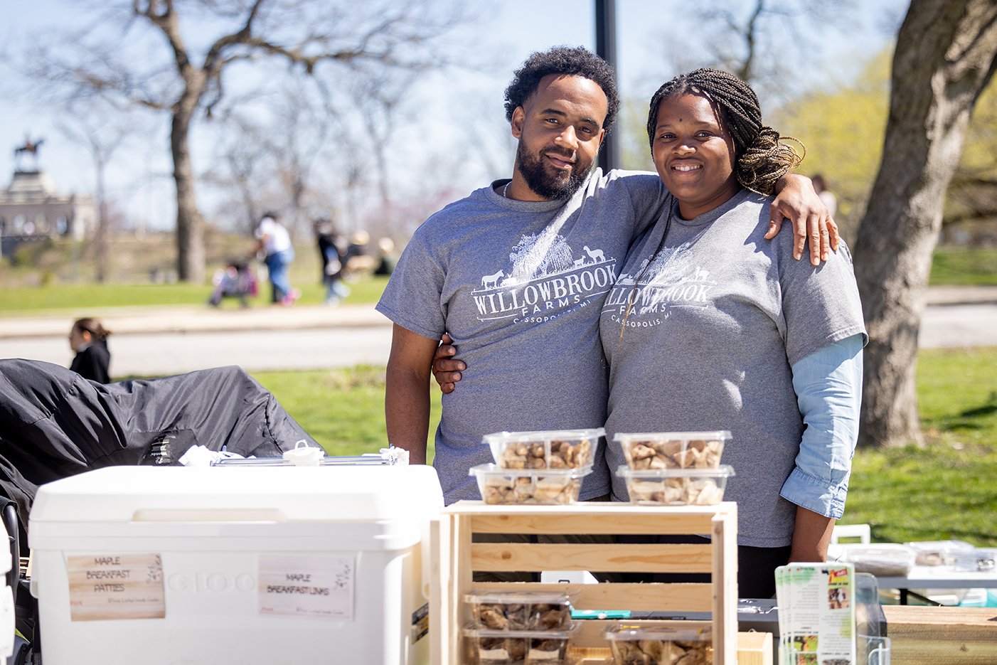 A man and woman pose with their arms around each other outside behind a table with their goods