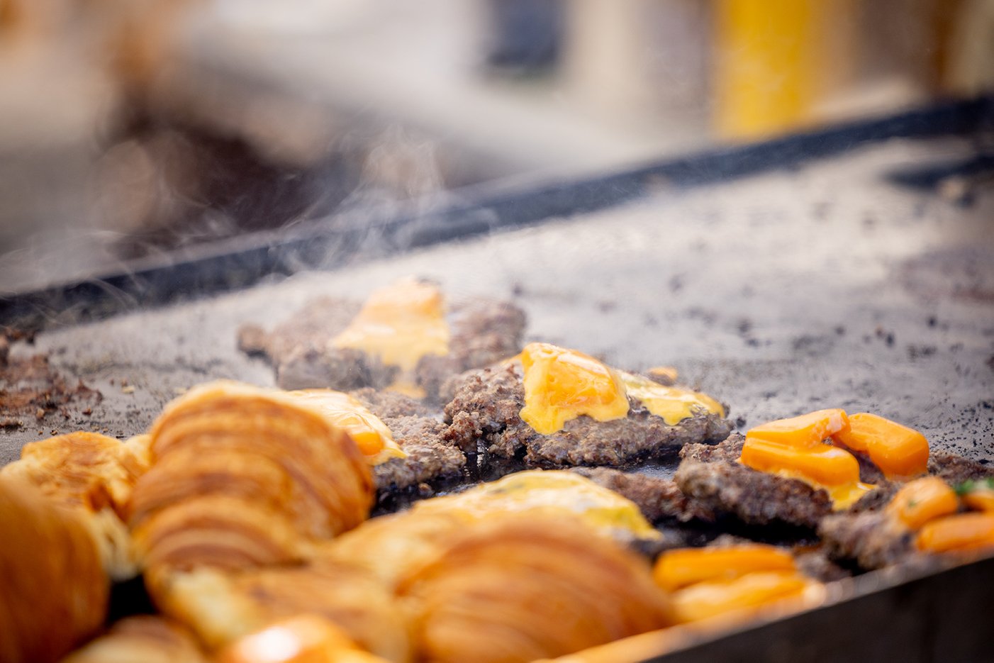Cheese-topped meat patties sizzling on a griddle next to croissants