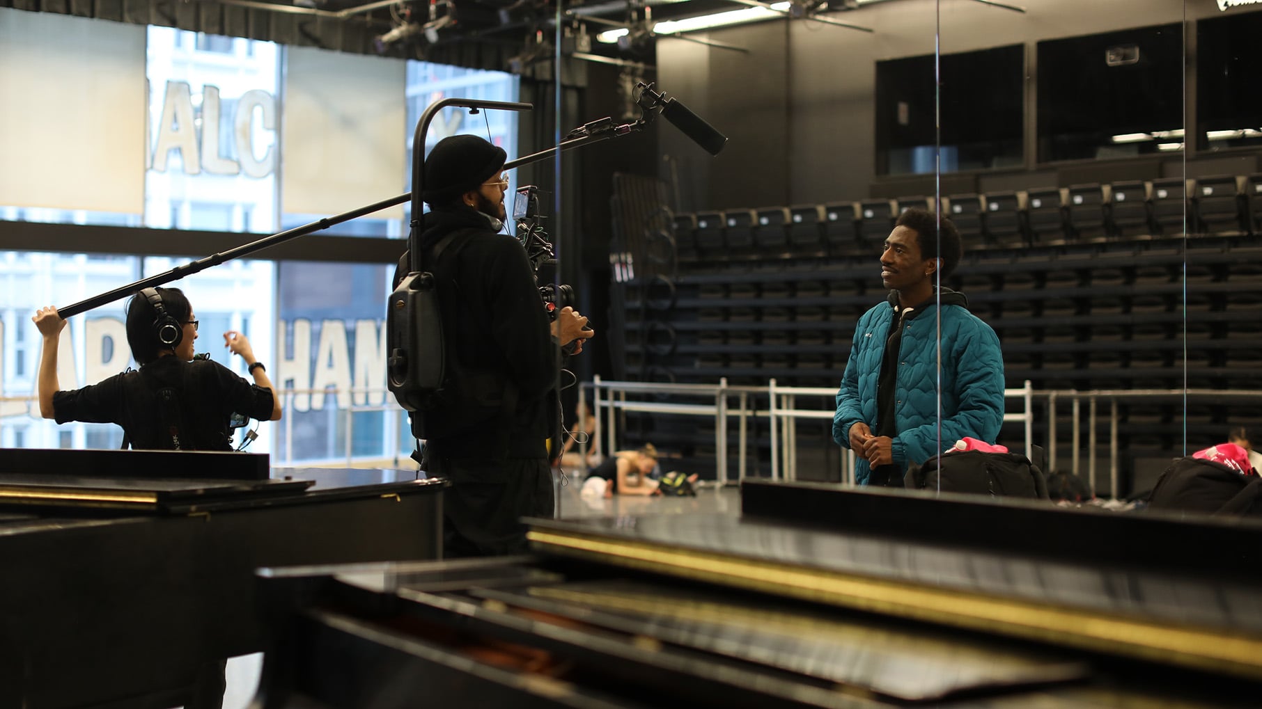 A Black man is interviewed behind a piano for a documentary by a man wearing a camera and a woman holding a boom
