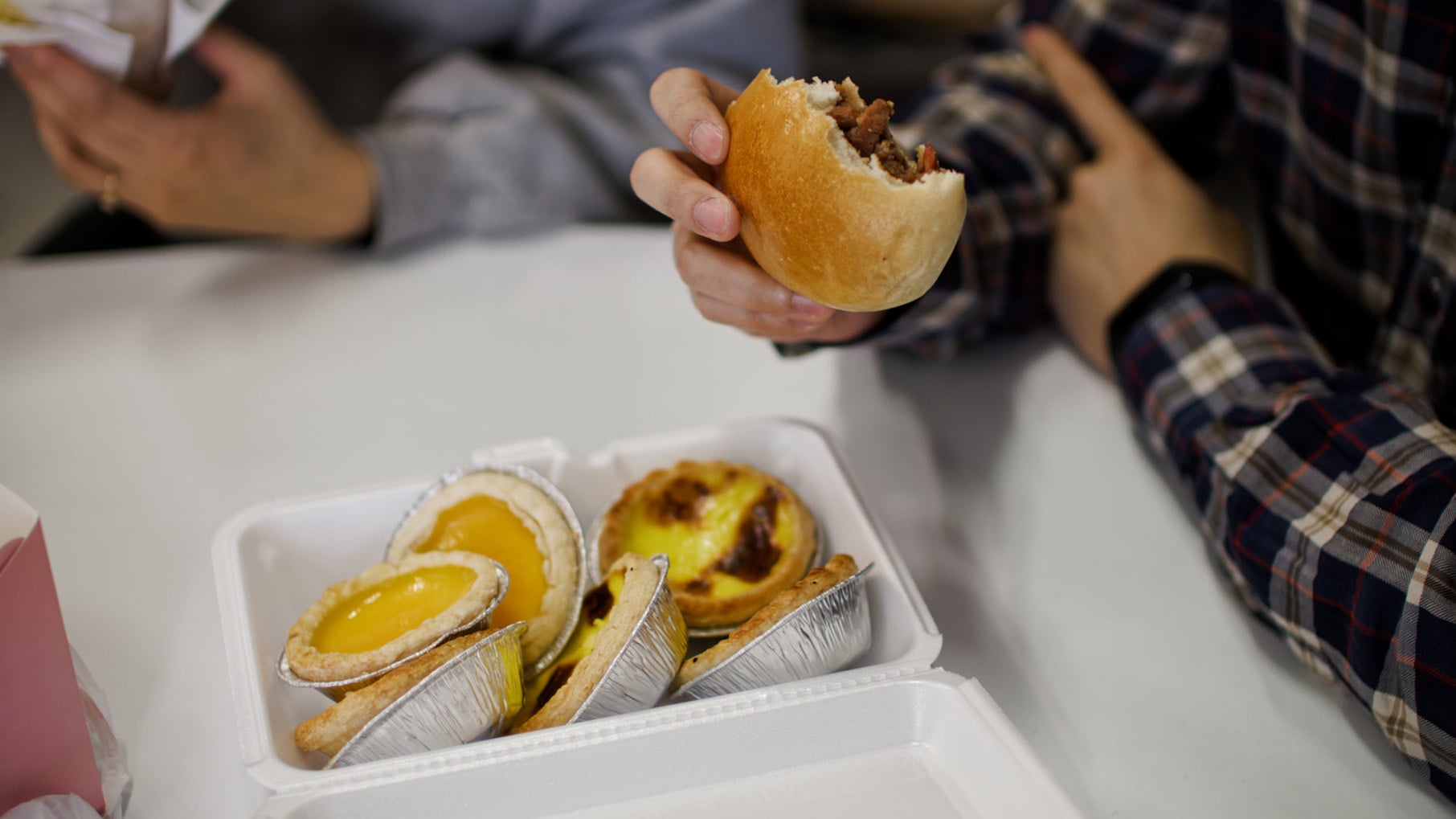 A hand holds a pork bun with a bite taken out of it over a styrofoam container of egg tarts