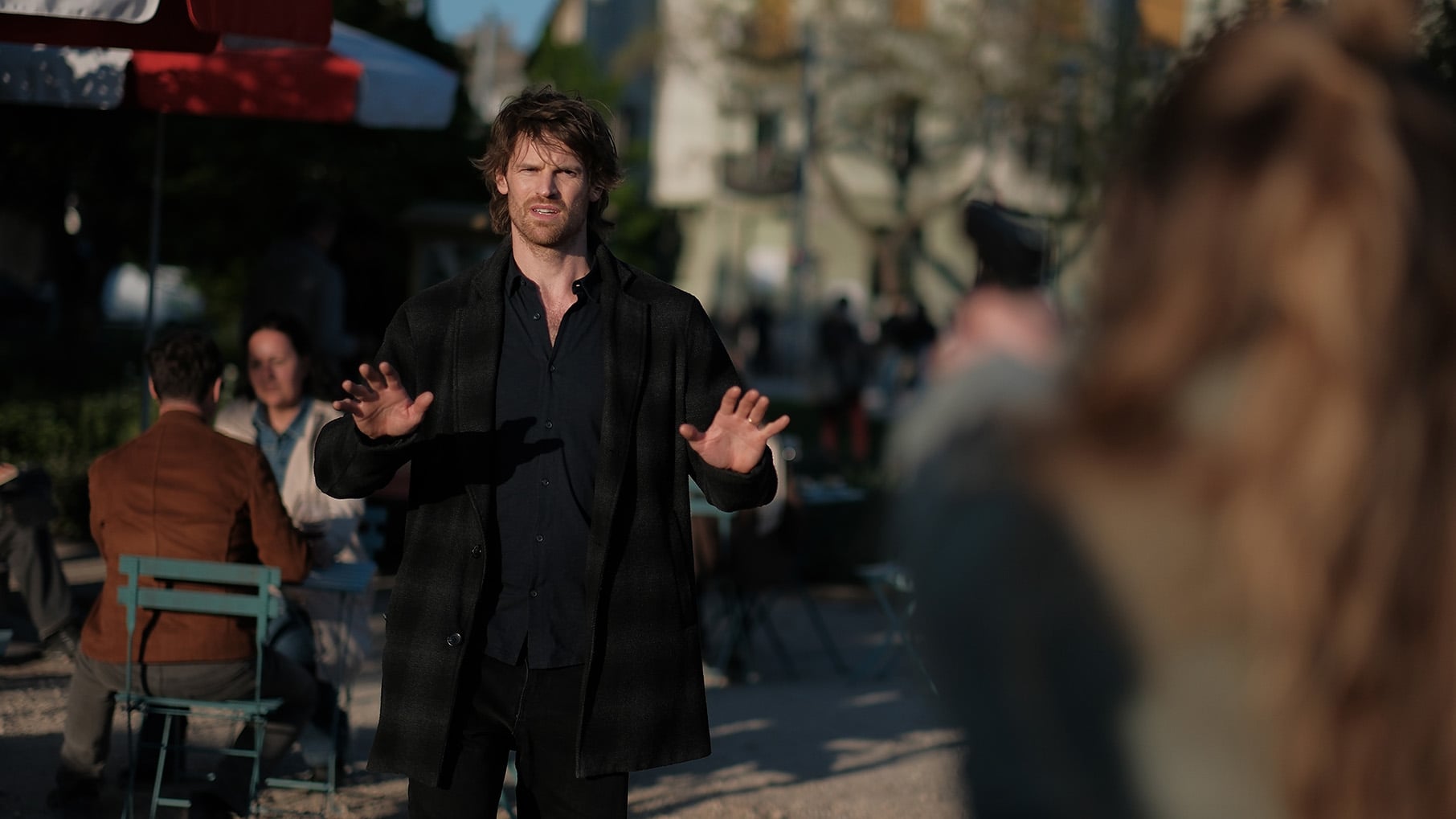 Maigret stands with his hands up in front of a table outside while a woman out of focus points a gun at him