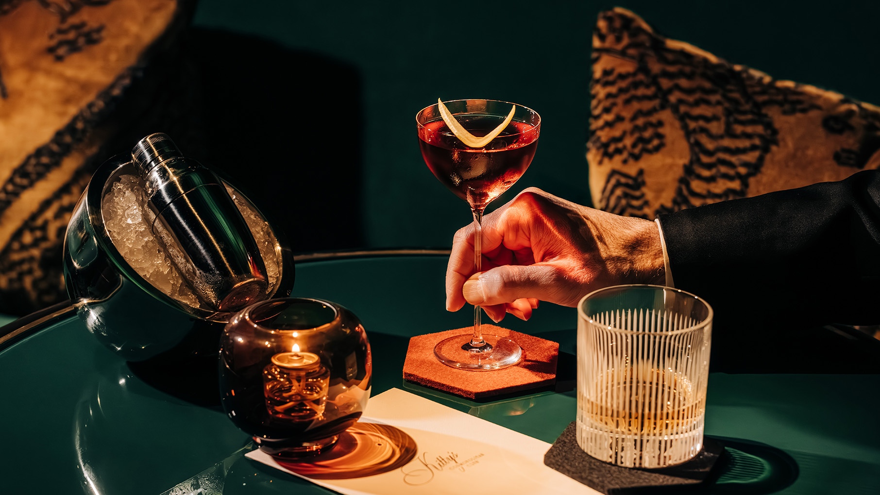 An arm reaches for a cocktail on a table with another glass and a candle in moody lighting