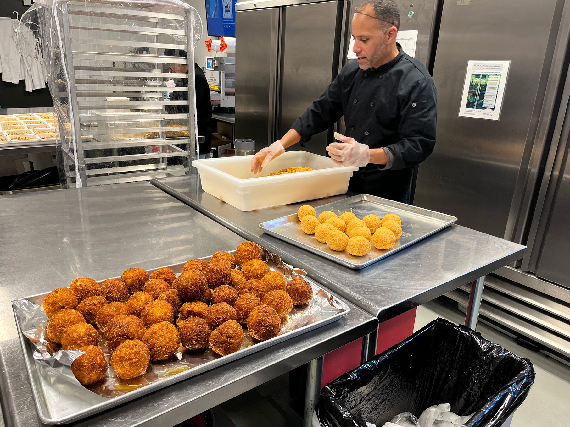 A man mixes rice in a tub in an industrial kitchen while a tray of fried rice balls sits in front of him