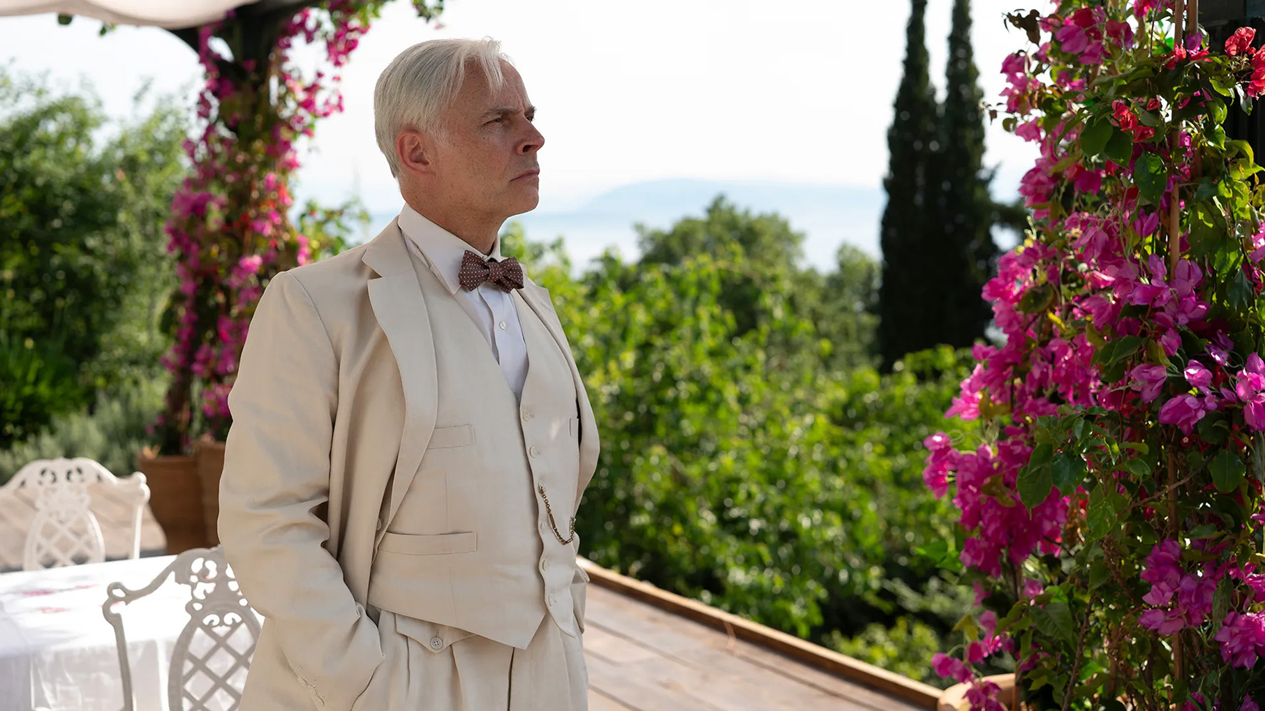 Mark Bonnar stands in a tan three-piece suit with a bowtie on a patio