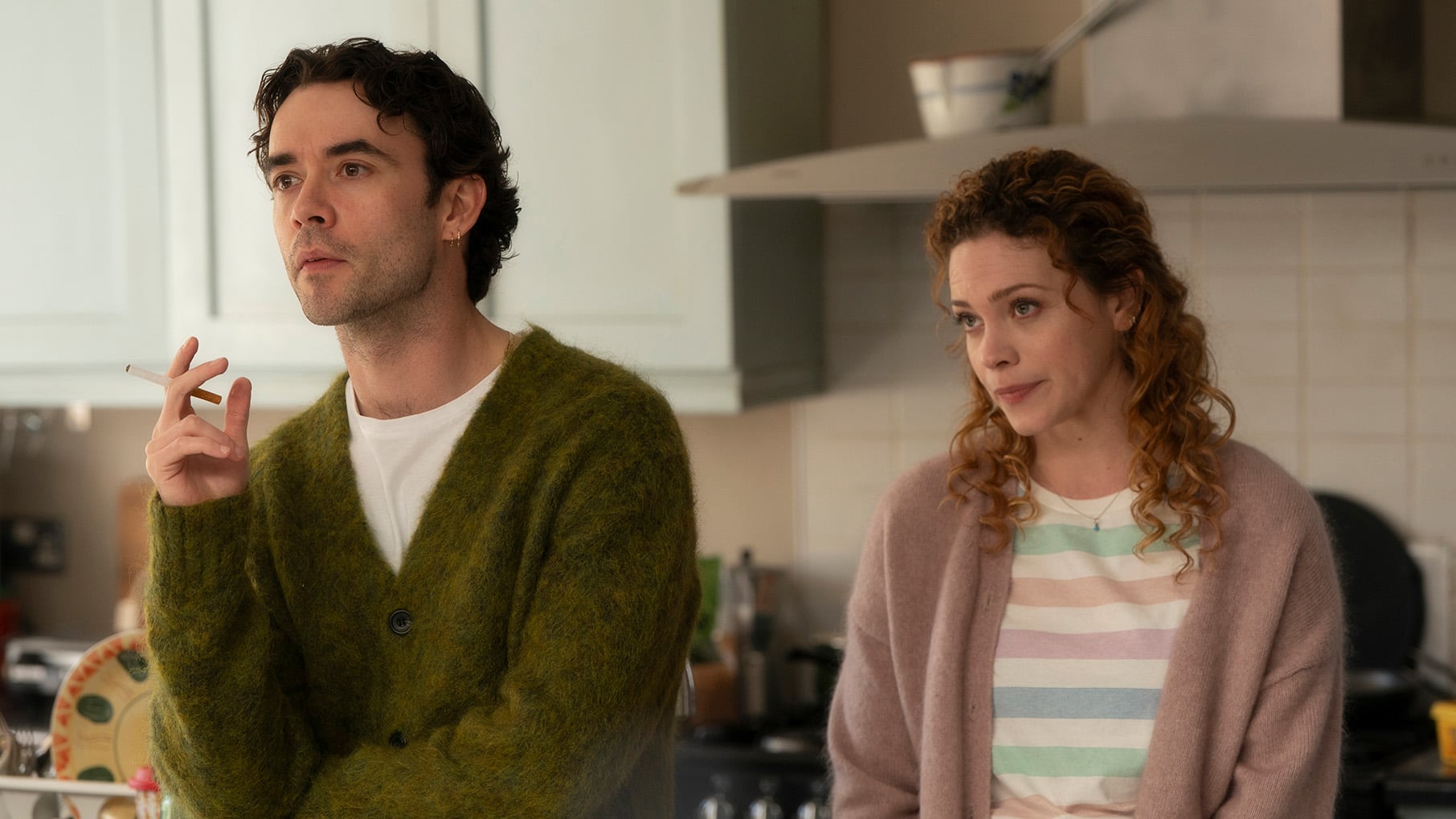 Elliot holds a cigarette next to Gillian in a kitchen