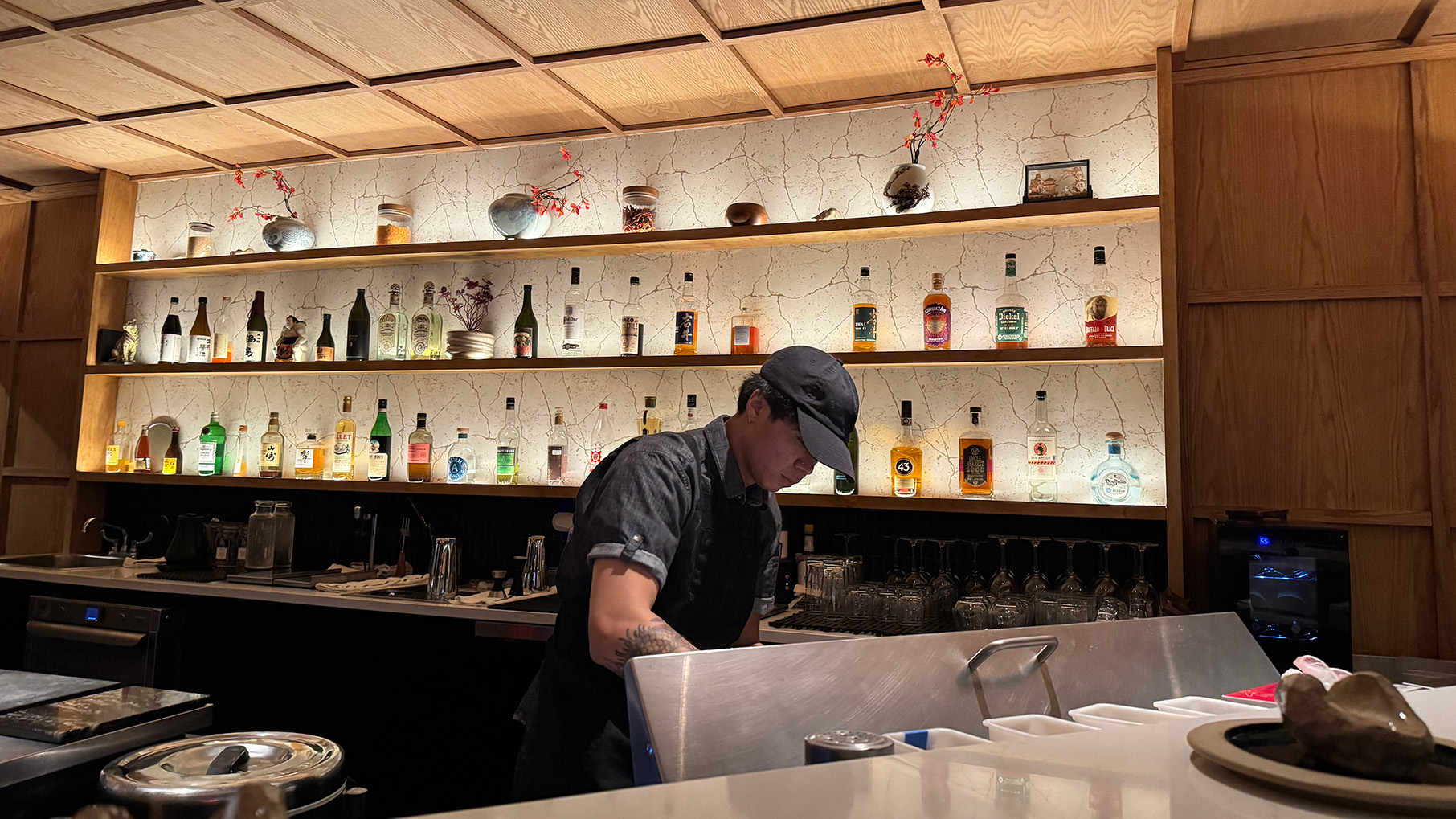 A man in a baseball cap behind a bar, with bottles on the wall behind him