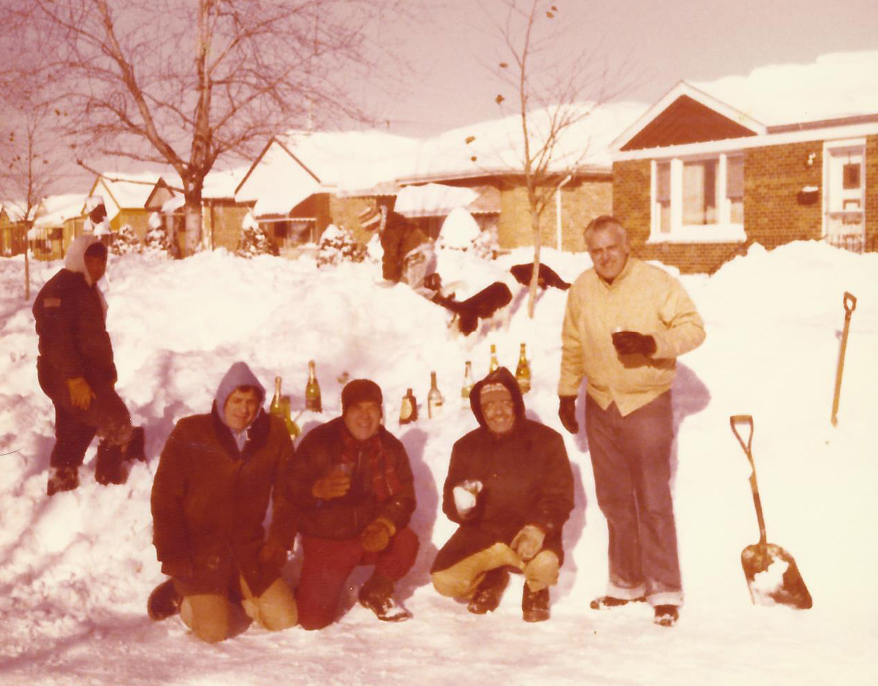 A group of men posing in front of a snow pile containing wine bottles stuck in the snow