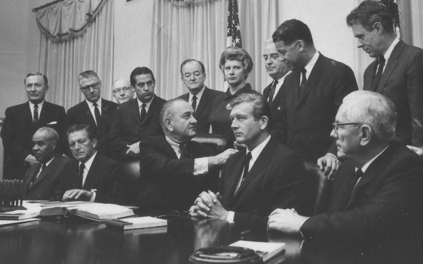 President Lyndon Johnson sits at a table surrounded by members of a commission in a black and white photo