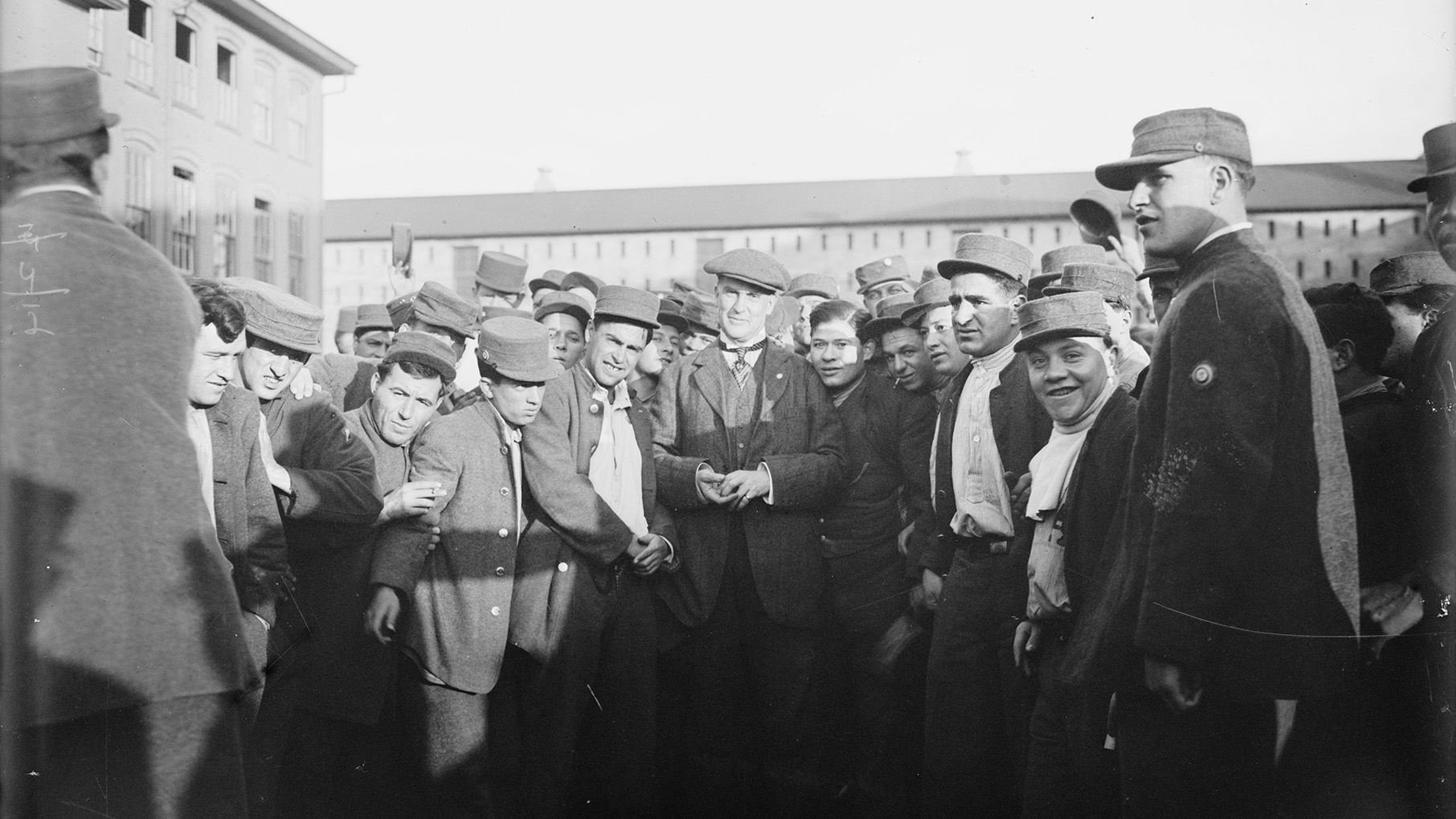 A black and white photo of men in caps standing outside a large building, some smiling for the camera