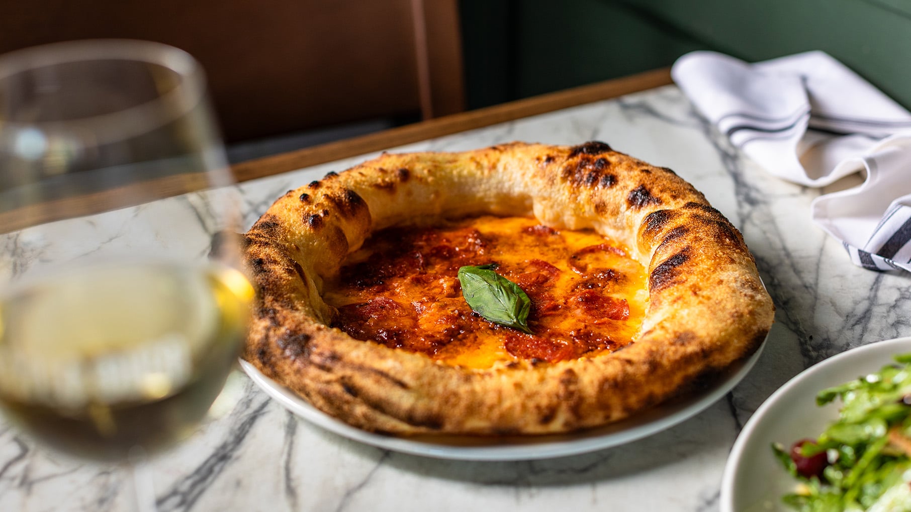 A pizza on a plate on a marble table with a glass of wine and salad