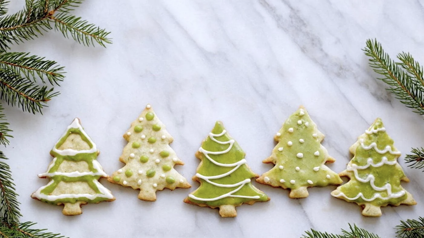 Christmas tree cookies decorated with green icing on a marble countertop, surrounded by spruce leaves
