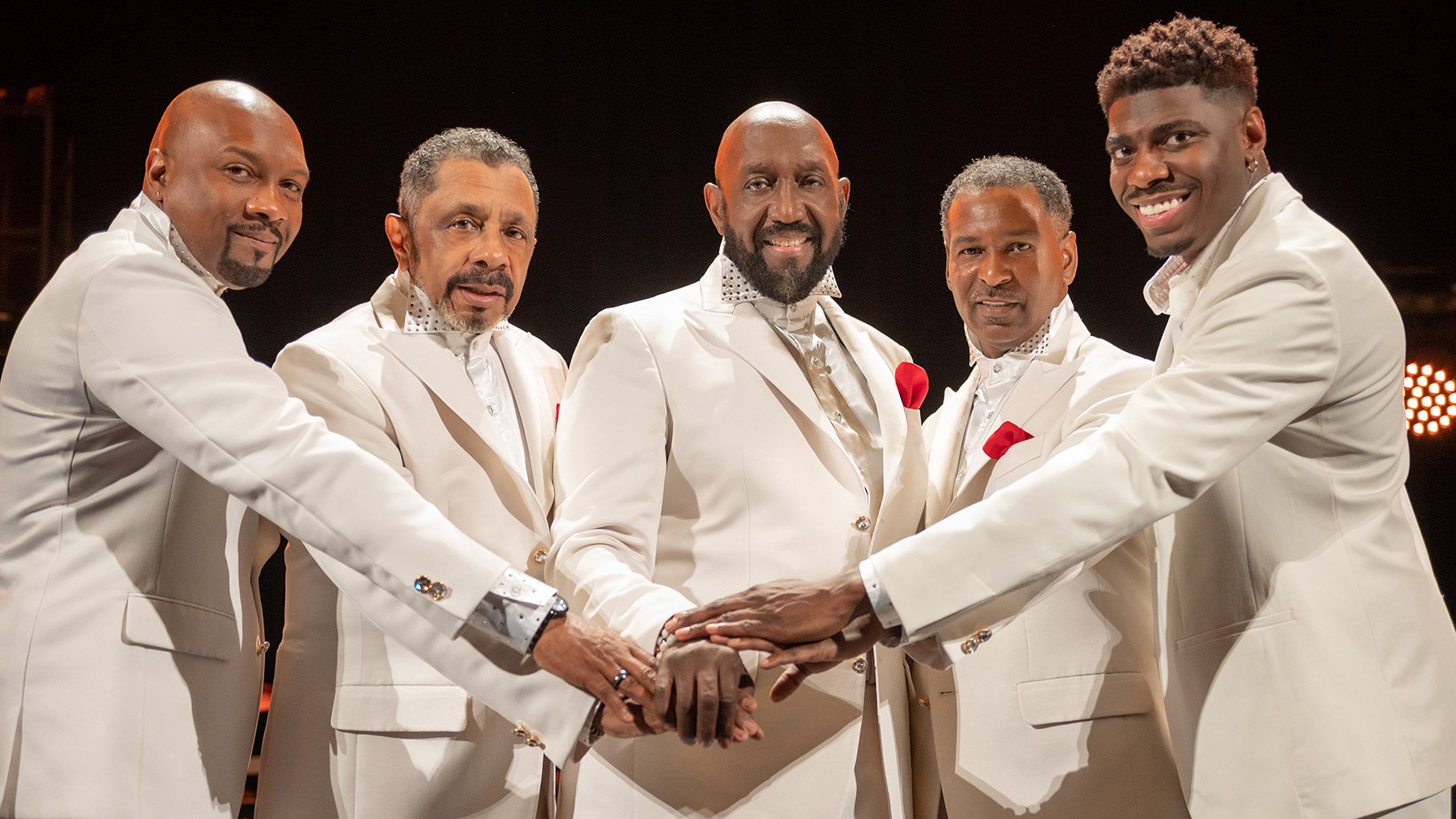 The Temptations, wearing white suits, smile and put their hands together