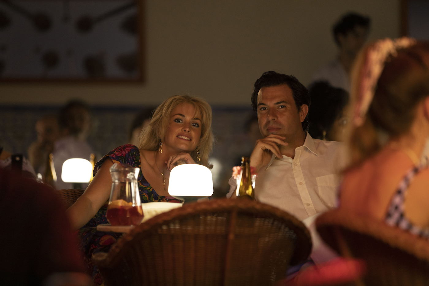 A woman and a man sit at a table amongst others with drinks on it and look at something 