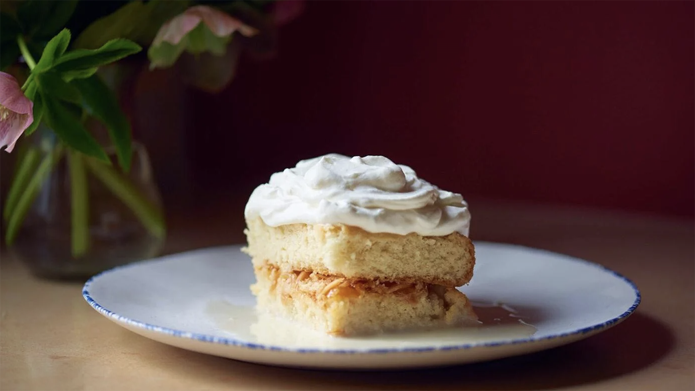 A slice of tres leches cake on a white plate with a vase of flowers in the background