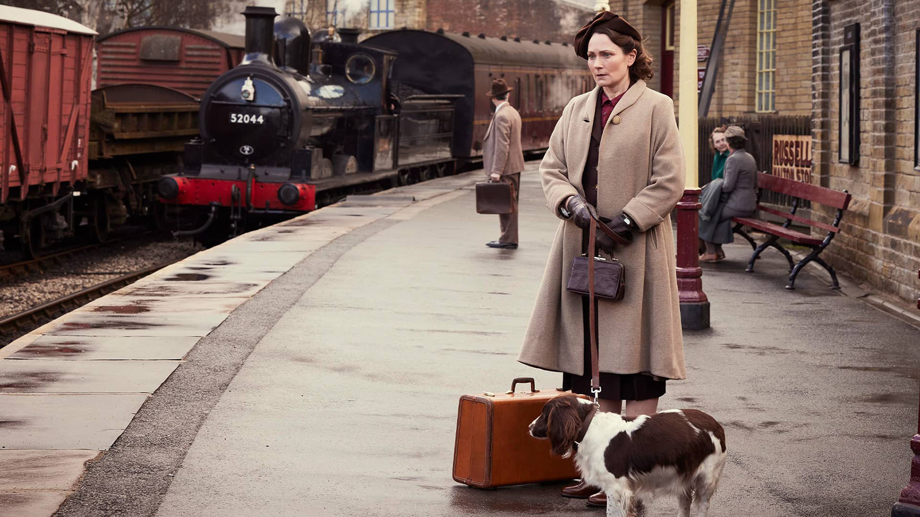 Mrs. Hall stands at a train station with a dog on a leash and luggage