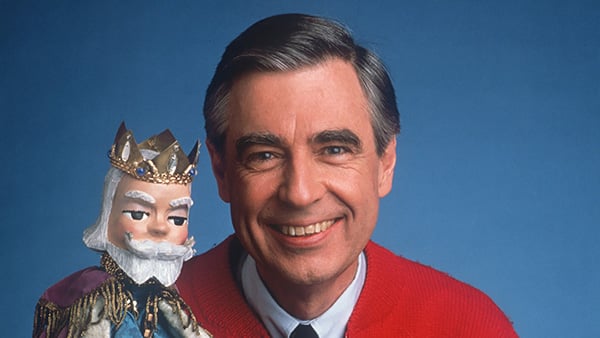 Mr. Rogers wearing his signature red cardigan, holding up the King Friday puppet against a blue backdrop
