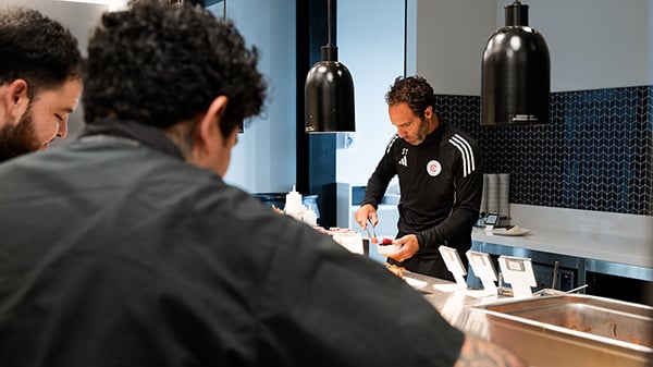 One Off Hospitality, the group behind The Publican and avec, runs the kitchen at the Chicago Fire's training facility. Credit: Jack X. Li for WTTW A man visible over another man's shoulder through a pass serves himself food
