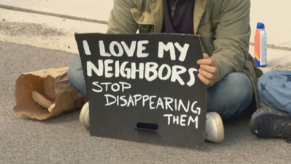 A person sitting cross-legged on a street with a sign reading "I love my neighbors. Stop disappearing them."