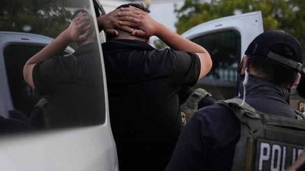 A man with his hands behind his head, facing away from the camera, is put into a van by a man wearing a tactical vest.