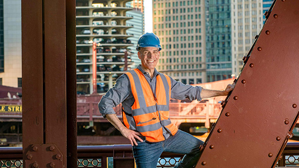 Geoffrey Baer poses in a hard hat and safety vest amongst the girders on a bridge