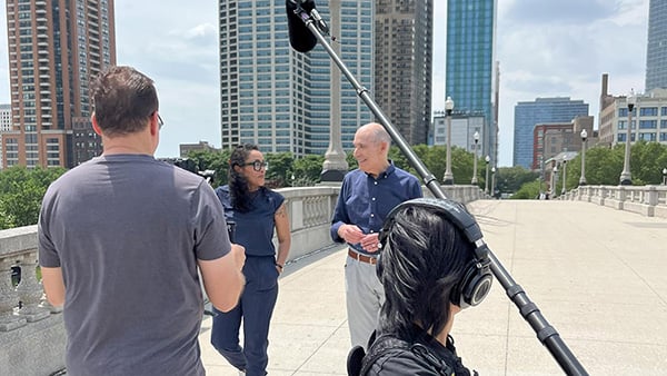 Geoffrey Baer walks with a woman under a microphone outside with a man filming