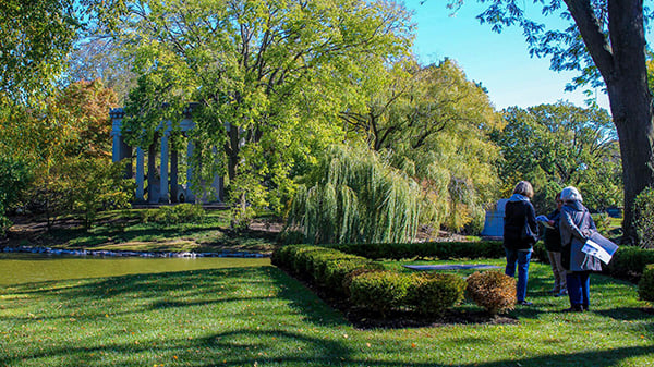 A small group of people stands in a cemetery with lots of trees, a stream, and a columned monument