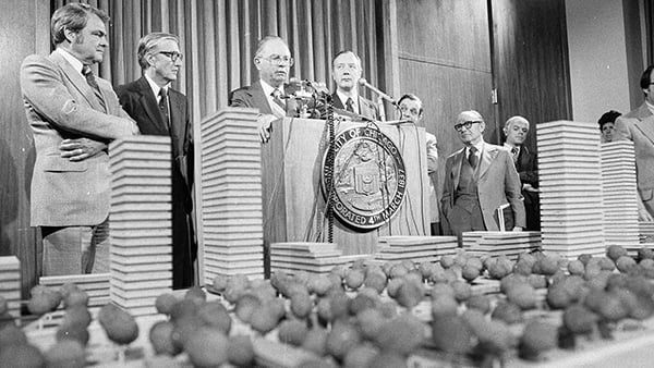 Men in suits stand on a stage with a podium behind a model of a neighborhood in a black and white photo