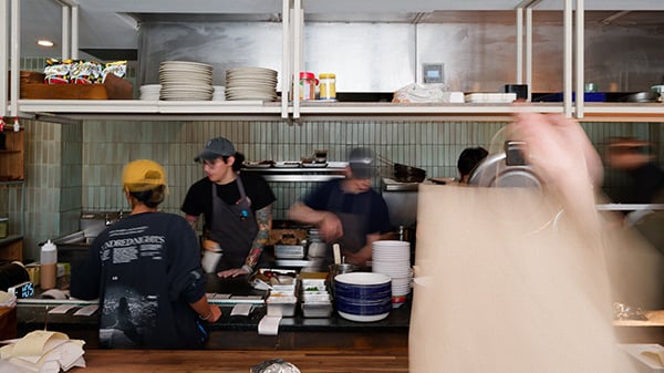 A hand lifts a paper bag in the foreground while people work in an open kitchen in the background