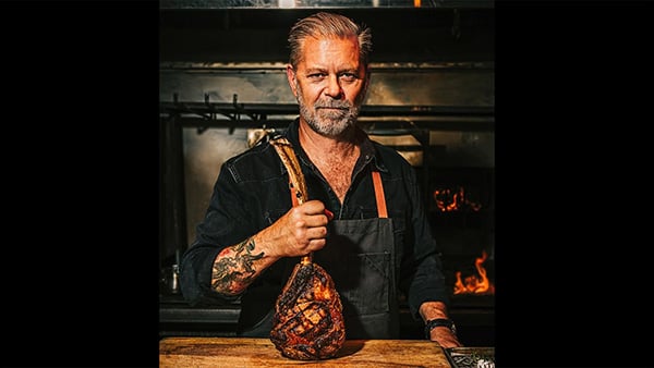 John Manion stands in a kitchen in front of a fire holding a steak by a bone