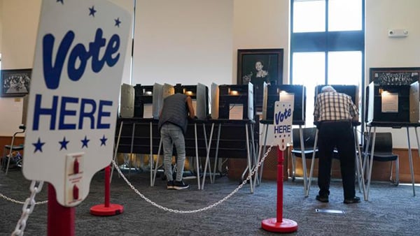 A sign reading "Vote Here" with people at voting booths in the background